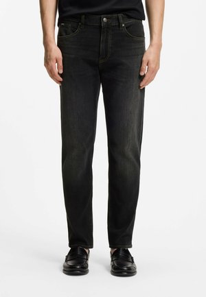 Man draagt strakke zwarte jeans en zwarte leren loafers, staand tegen een effen witte achtergrond.
