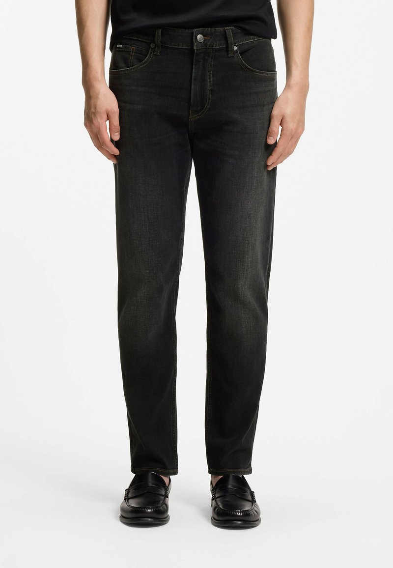 Homme portant un jean noir slim et des mocassins en cuir noir, debout devant un fond blanc uni.