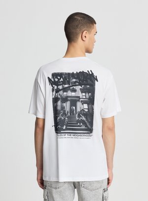 Giovane uomo visto di spalle che indossa una maglietta bianca con una foto in bianco e nero di persone sedute su una scala urbana e testo con graffiti.