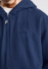 Námornícka modrá mikina na zips vyrobená z bavlny, s jemnou textúrou, kapucňou a vyšívaným logom na ľavej hrudi. Hladká zipsová súčiastka.