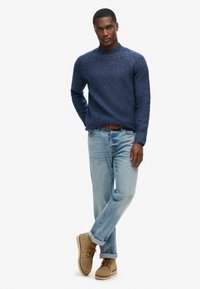 Marineblauwe gebreide trui met ronde hals en geribbelde manchetten, gecombineerd met lichtblauwe jeans en tan kleurige veterschoenen.