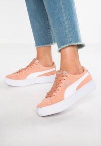 Sneakers Puma w jasnoróżowym zamszu z białymi akcentami, okrągłym noskiem, płaską białą podeszwą i teksturowanymi sznurówkami. Ubrane z podwiniętymi niebieskimi dżinsami.