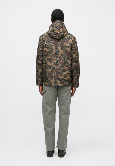 Vīrietis stāv ar muguru pret skatītāju, tērpies brūnā un zaļā maskēšanās jaka ar kapuci, pelēnās kravas biksēs un tumši brūnās zābakos uz baltas virsmas.