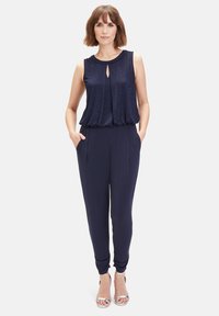 Marineblauer Jumpsuit mit einem gefalteten Oberteil, Schlitzdetail, schmal zulaufenden Beinen und seitlichen Taschen. Der Stoff hat eine glatte Textur und einen leichten Glanz. Silberne Absätze.