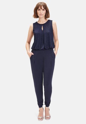 Combinaison bleu marine avec un haut plissé, un détail en ouverture clé, des jambes fuselées et des poches latérales. Le tissu a une texture lisse et un léger éclat. Talons argentés.