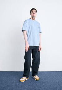 Light blue cotton t-shirt, relaxed fit, with short sleeves and a small yellow logo. Paired with dark pants and orange-white sneakers.