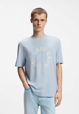 Junger Mann mit kurzen Haaren, der ein hellblaues T-Shirt mit dem Schriftzug "LAID BACK LIFE" im Kreis und hellblaue Jeans trägt, steht mit einer Hand in der Tasche.