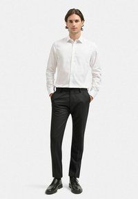 Vit långärmad skjorta med knappar, matchad med skräddarsydda svarta byxor och svarta snörskor, presenterad på en manlig modell.