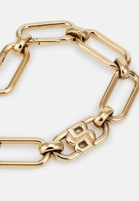 Brățară din metal auriu cu link-uri elongate și un sistem de închidere proeminent ce include un logo stilizat. Suprafața este lucioasă și netedă.