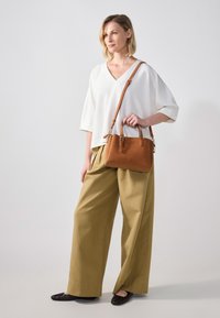 Borsa in pelle marrone con doppio manico e tracolla regolabile, indossata con una maglietta bianca a scollo a V e pantaloni beige a gamba larga. Scarpe scure e piatte.