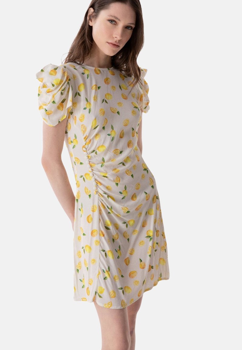 Junge Frau in einem weißen Kleid mit gelben Zitronenmustern und gepufften kurzen Ärmeln, stehend vor einem schlichten Hintergrund.