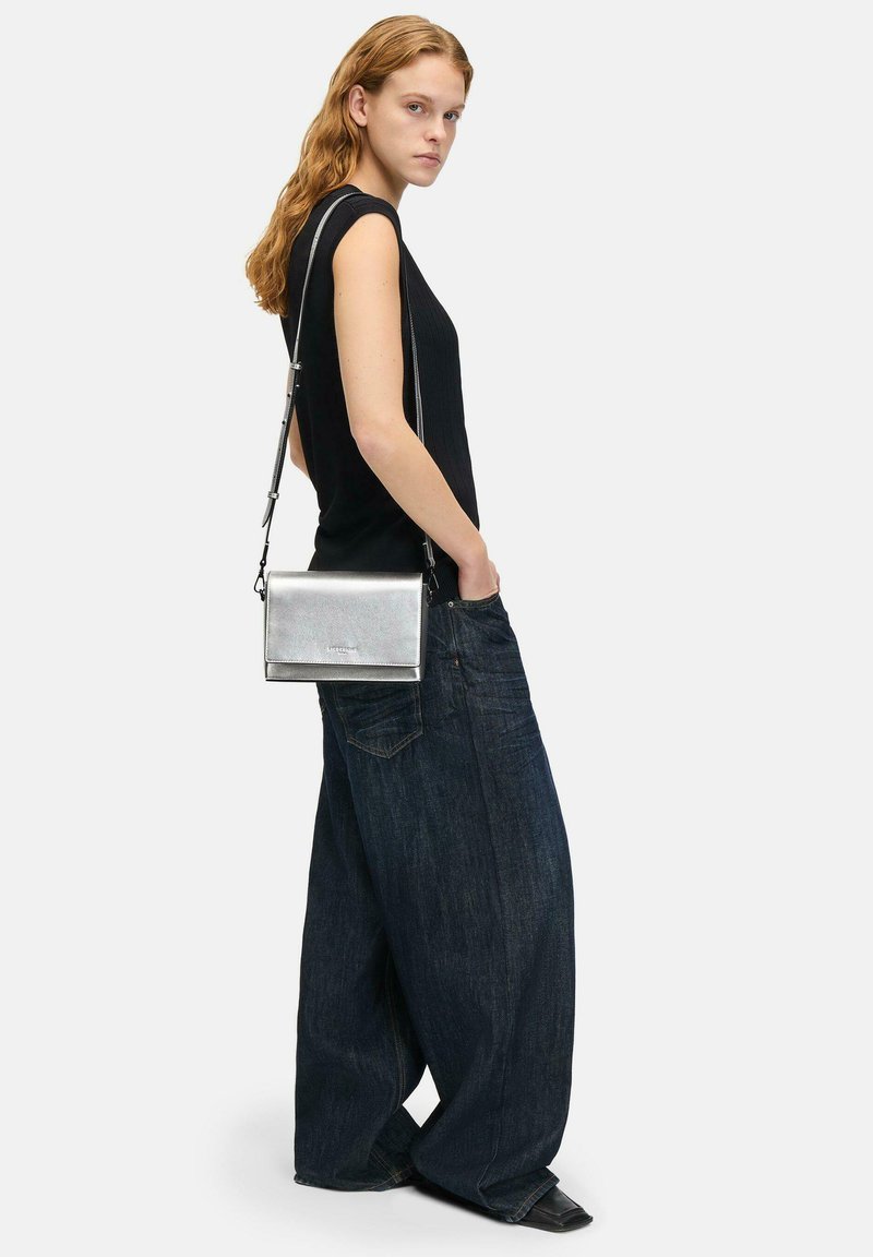 Silberne metallische Umhängetasche in rechteckiger Form, mit glatter Textur und verstellbarem Schultergurt, getragen mit dunkelblauen, oversized Jeans.