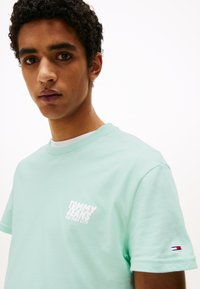 Menta zöld pamut póló, fehér "TOMMY JEANS NEW YORK CITY" logóval a mellkasán és egy kis háromszínű logóval az ujjon.