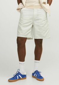 Pantaloni corto di colore chiaro, lunghi fino al ginocchio, realizzati in tessuto misto cotone con una texture liscia. Abbinati a scarpe da ginnastica blu e calze bianche.