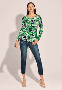 Langärmliges Oberteil in Grün und Marineblau mit wirbelnden Mustern und Text. Kombiniert mit dunkelblauen engen Jeans und weißen Absätzen.
