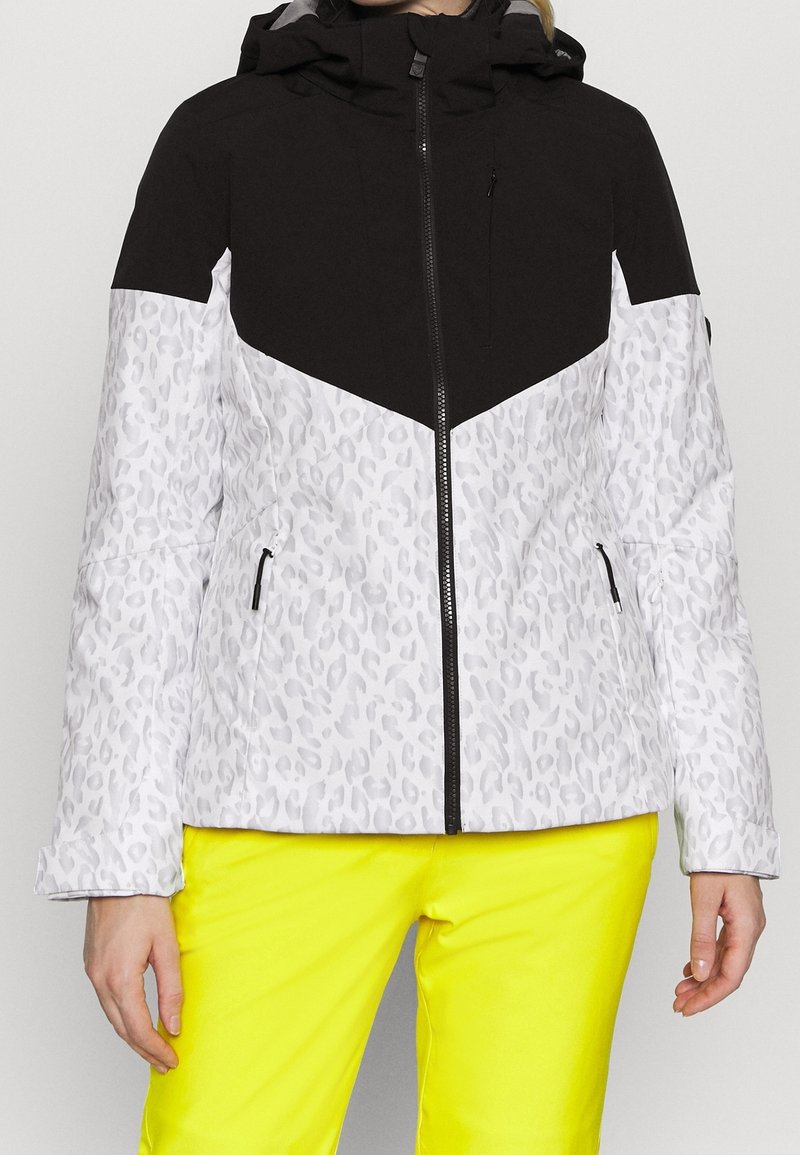 Veste à capuche imprimé léopard noir et blanc avec fermeture éclair noire et poches, portée avec un pantalon jaune vif.
