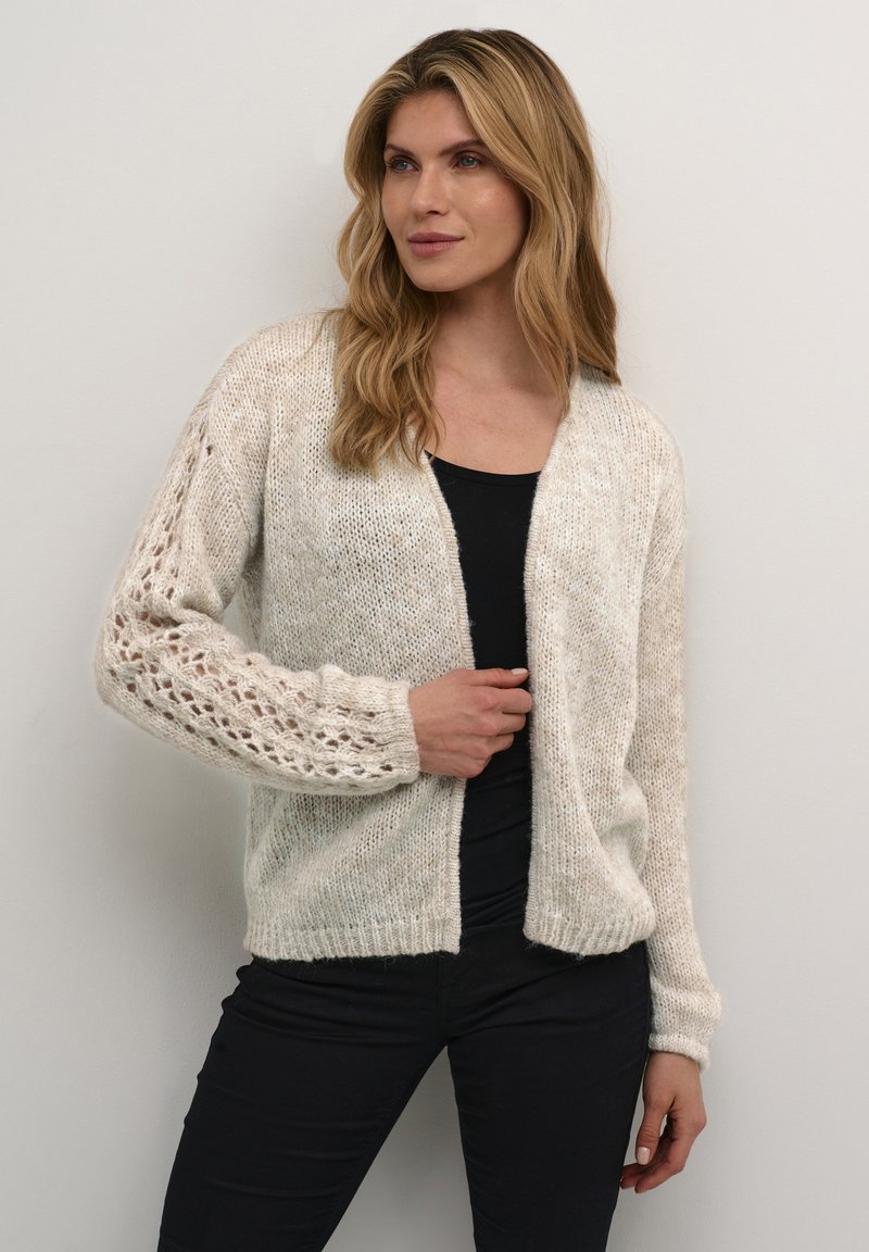 Light beige cardigan with a loose knit texture, featuring a lace-patterned sleeve and ribbed cuffs. Worn over a black top.