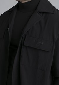 Svart lättviktsjacka med dragkedja, framficka med präglad logotyp och krage. Burits över en svart polotröja. Slät textur.