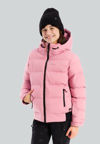 Niña sonriente con un gorro negro y una chaqueta acolchada rosa con bolsillos con cremallera, de pie frente a un fondo gris claro.