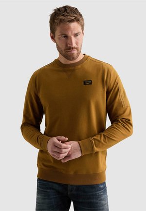 Skæggede mand iført en sennepsgul sweatshirt med en lille brystlomme, står med hænderne foldet og kigger til siden.