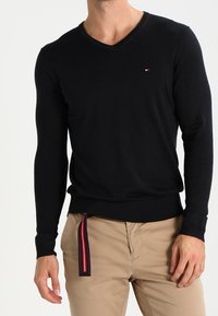 Czarny sweter typu v-neck z miękkiego materiału, z długimi rękawami i subtelnym logo na piersi. Noszony na beżowych spodniach.