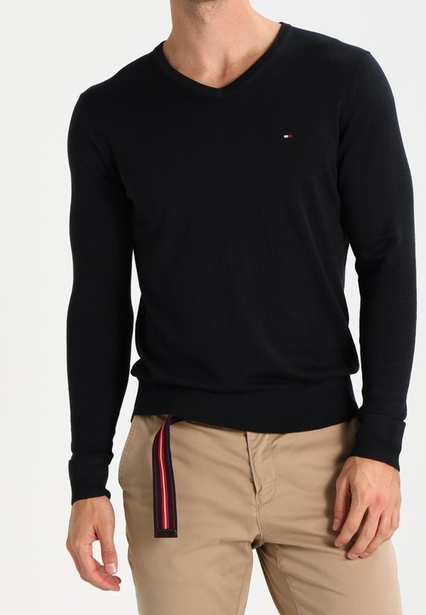 Czarny sweter typu v-neck z miękkiego materiału, z długimi rękawami i subtelnym logo na piersi. Noszony na beżowych spodniach.