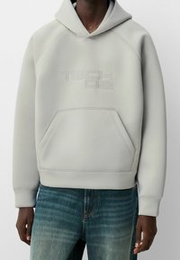 Ljusgrå hoodie i slät tyg, med en känguruficka och präglad "TECH 02"-logotyp på framsidan. Huvan och ribbade ärmslut.