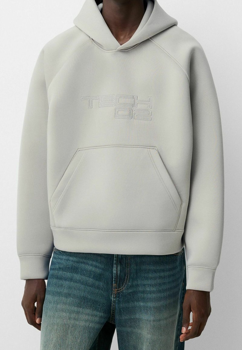Ljusgrå hoodie i slät tyg, med en känguruficka och präglad "TECH 02"-logotyp på framsidan. Huvan och ribbade ärmslut.