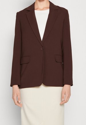 Kobieta ubrana w ciemnobrązową marynarkę na jeden guzik, na białym topie i kremowej spódnicy, stojąca na jednolitym białym tle.