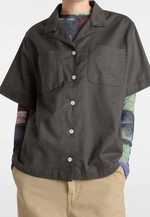 Personne portant une chemise à manches courtes gris foncé boutonnée avec deux poches poitrine par-dessus un haut coloré à manches longues et un pantalon beige.