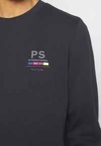 Sweat bleu marine avec le texte « PS International », barres colorées en bleu, rose et jaune, et petits noms de villes en dessous sur la poitrine.