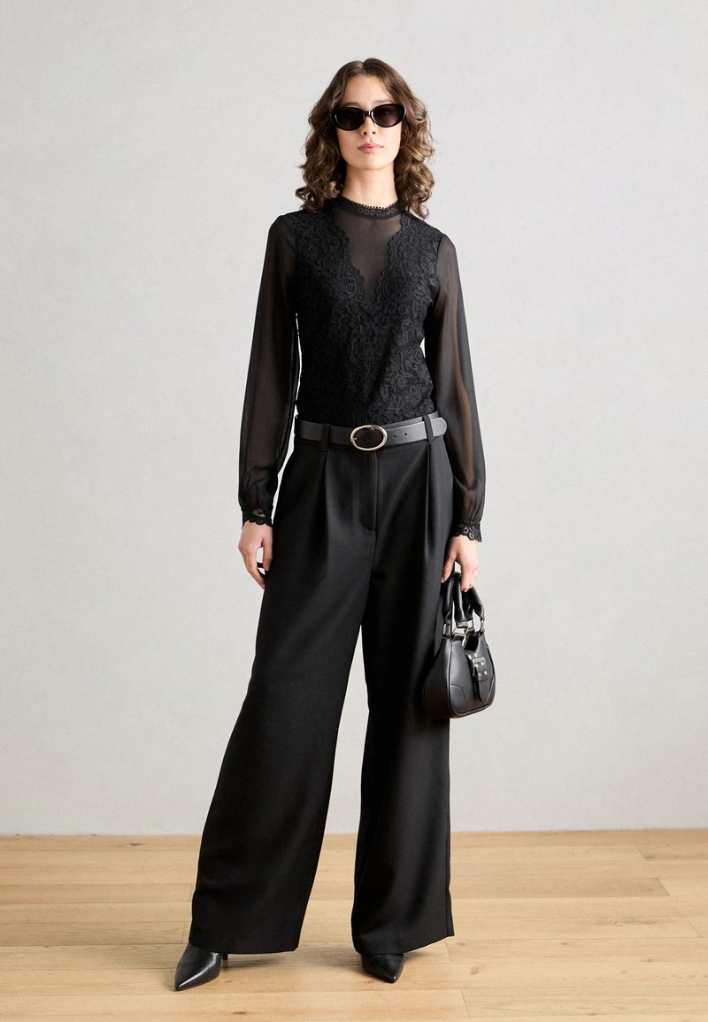 Blusa negra transparente con detalles de encaje, pantalones anchos negros y tacones negros puntiagudos. Los accesorios incluyen un bolso negro estructurado y gafas de sol.