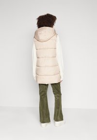 Světlé béžové péřové vesta s kapucí, s vertikálním prošíváním, v kombinaci s olivově zelenými kordovými kalhotami a bílými platformovými teniskami.
