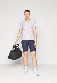 Camicetta viola chiaro a maniche corte, pantaloni ciclistici blu navy, mentre tiene una borsa a tracolla nera. Scarpe e calze bianche, in piedi contro uno sfondo chiaro.