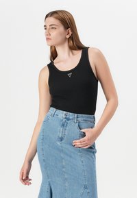 Model poseert zelfverzekerd, gekleed in een zwarte tanktop en een high-waisted spijkerrok, met een schone, witte achtergrond.
