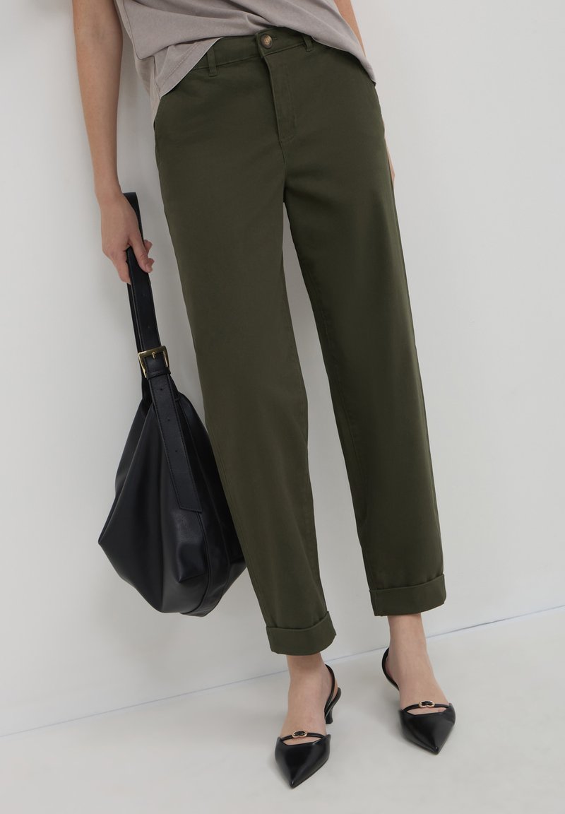 Pantaloni a sigaretta verde oliva con orli risvoltati, abbinati a scarpe slingback nere a punta e a una grande borsa a tracolla in pelle nera.