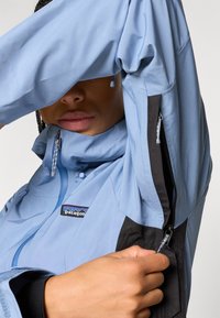 Ανοιχτό μπλε μπουφάν με μαύρες λεπτομέρειες, που διαθέτει ρυθμιζόμενα κορδόνια, φερμουάρ και λογότυπο. Λείο, αδιάβροχο υλικό.