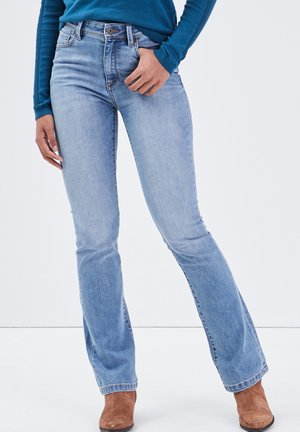 Vrouw draagt lichtblauwe bootcut jeans, een teal geribbelde trui en bruine suède enkellaarsjes, staand met handen bij de zakken.