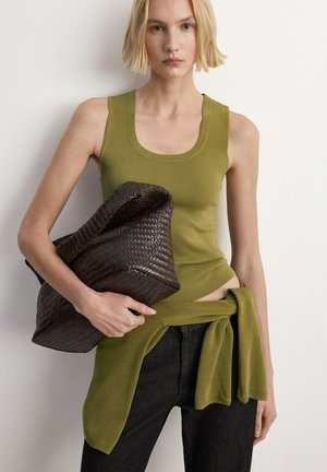 Žena s krátkými blond vlasy, oblečená v olivově zeleném tílku a svetru přivázaném v pase, držící velké tmavé pletené psaníčko.