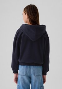 Kvinna med långt brunt hår som bär en marinblå fleecehoodie och ljusblå jeans, sedd bakifrån mot en enkel grå bakgrund.