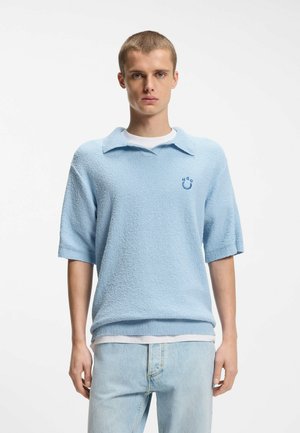 Junger Mann trägt ein hellblaues, strukturiertes Poloshirt mit kurzen Ärmeln und Kragen sowie hellblaue Jeans, steht vor einem schlichten weißen Hintergrund.