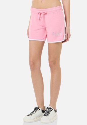 Frau trägt pinke Shorts mit Kordelzug und weißem Saum sowie weiße Sneaker mit schwarzen Details, steht vor einem einfarbigen Hintergrund.