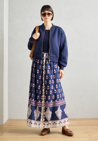 Veste bomber zippée marine, haut sombre, jupe maxi florale en marine avec des motifs rouges et blancs, mocassins marron, sac marron, lunettes de soleil oversize.