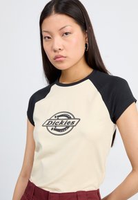 Eine Frau mit langen dunklen Haaren trägt ein kurzärmliges, beiges und schwarzes Shirt mit einem runden grafischen Logo. Sie hat einen neutralen Gesichtsausdruck.