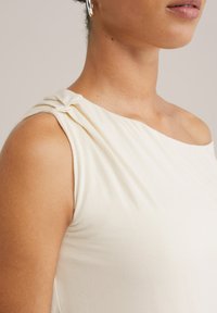 Top u boji kreme bez rukava s drapiranim dizajnom i naboranim detaljem na lijevom ramenu. Glatka tekstura i lagani materijal.