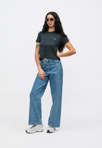 Svart crop t-skjorte med logo, lyseblå vide jeans, grå sneakers og lyse solbriller. Stående modell mot en hvit bakgrunn.