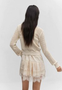 Suéter de punto beige con mangas largas, combinado con una falda corta de capas de tejido transparente que presenta un dobladillo de encaje con forma de ondas. Diseño simple y texturizado.