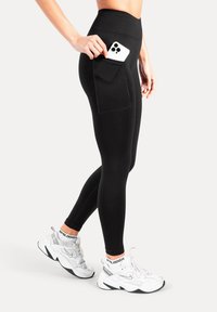 Leggings noirs taille haute avec poche latérale, tissu extensible, texture lisse et design élégant. Associés à des chaussures de sport blanches.