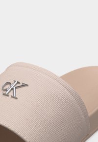 Sandale beige en tissu avec un logo "CK" métallique argenté sur la large bride et une semelle beige.