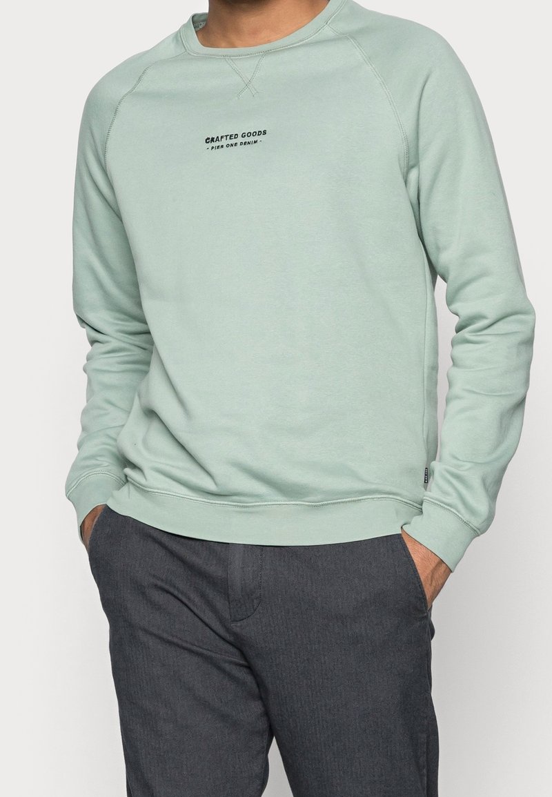 Sweat-shirt vert menthe à manches longues, poignets côtelés et col rond. Inscription "CRAFTED GOODS" en noir sur le devant. Porté avec un pantalon gris foncé.